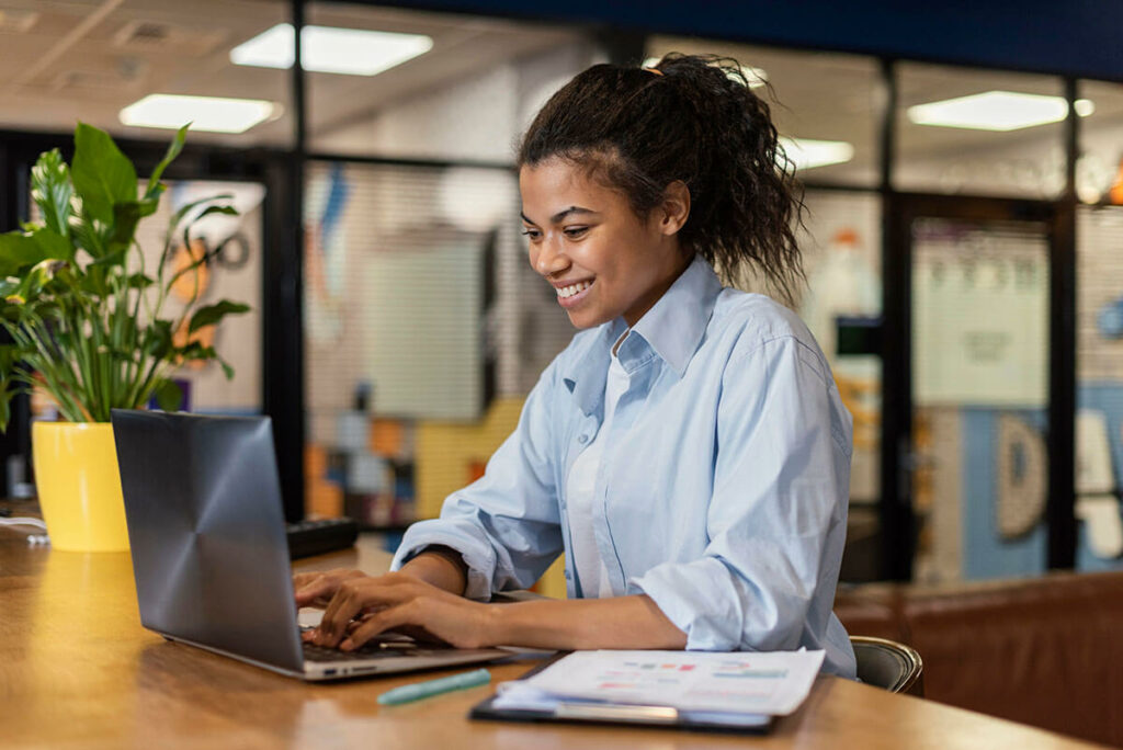 Uma mulher sorridente está trabalhando em um laptop em um ambiente de escritório moderno. Ela está sentada em uma mesa de madeira com uma planta em vaso amarelo ao lado. O fundo mostra um ambiente de trabalho com divisórias de vidro e iluminação clara.