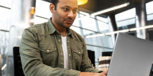 Homem usando uma camisa verde, concentrado em seu trabalho no laptop em um ambiente de escritório moderno e iluminado.