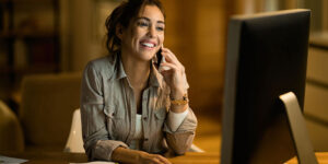 Uma jovem mulher, sorridente, fala ao telefone enquanto trabalha em um computador desktop à noite em casa. Ela está sentada em uma mesa com um bloco de notas, um lápis e uma xícara de café ao lado do teclado.